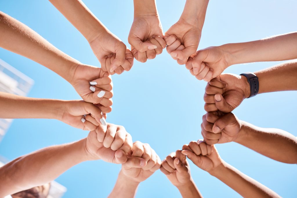 Various hands of different skin tones joining together to form a circle against a clear blue sky.