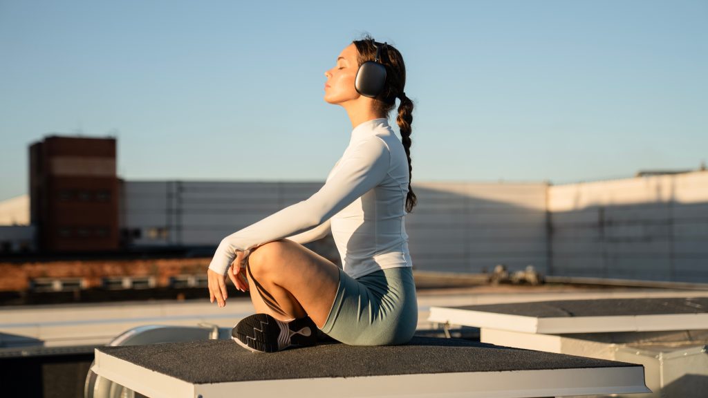 Young woman in a white long-sleeve top and blue shorts practicing lotus asana while wearing large headphones.
