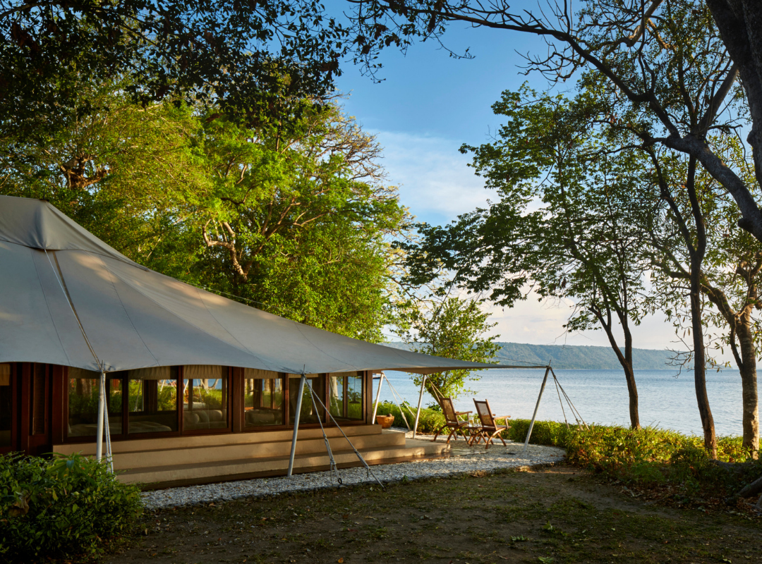 Canvas tent structure with large windows situated near a body of water surrounded by trees.