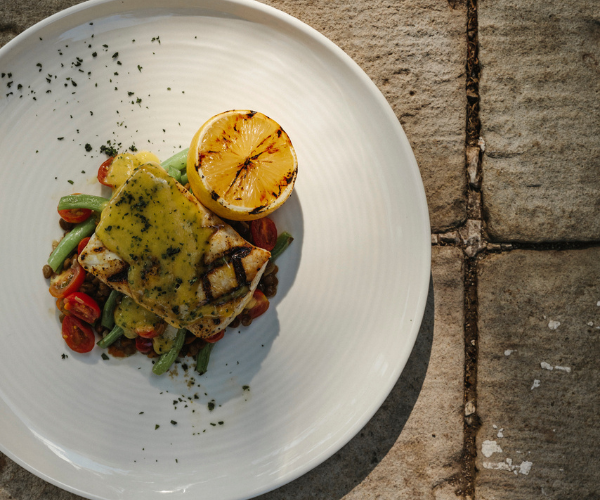 Plate of grilled fish topped with sauce, served with green beans, cherry tomatoes, and a grilled lemon half.