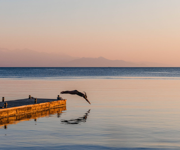 Individual diving from a wooden pier into still water during sunset, with a reflection visible on the surface.