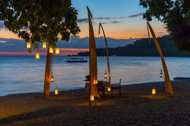 Lanterns hanging from tall poles on a beach during sunset, with a calm sea in the background.