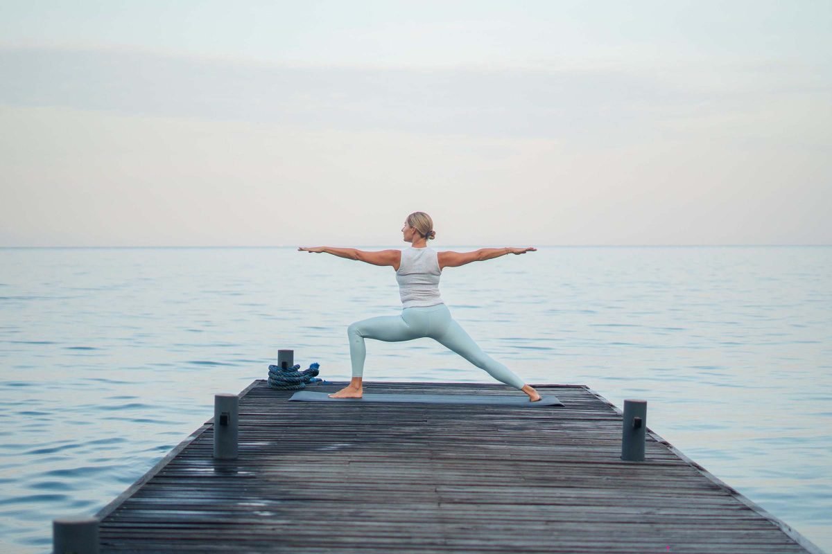 Woman in light blue yoga attire performing a warrior pose on a wooden dock over calm water.