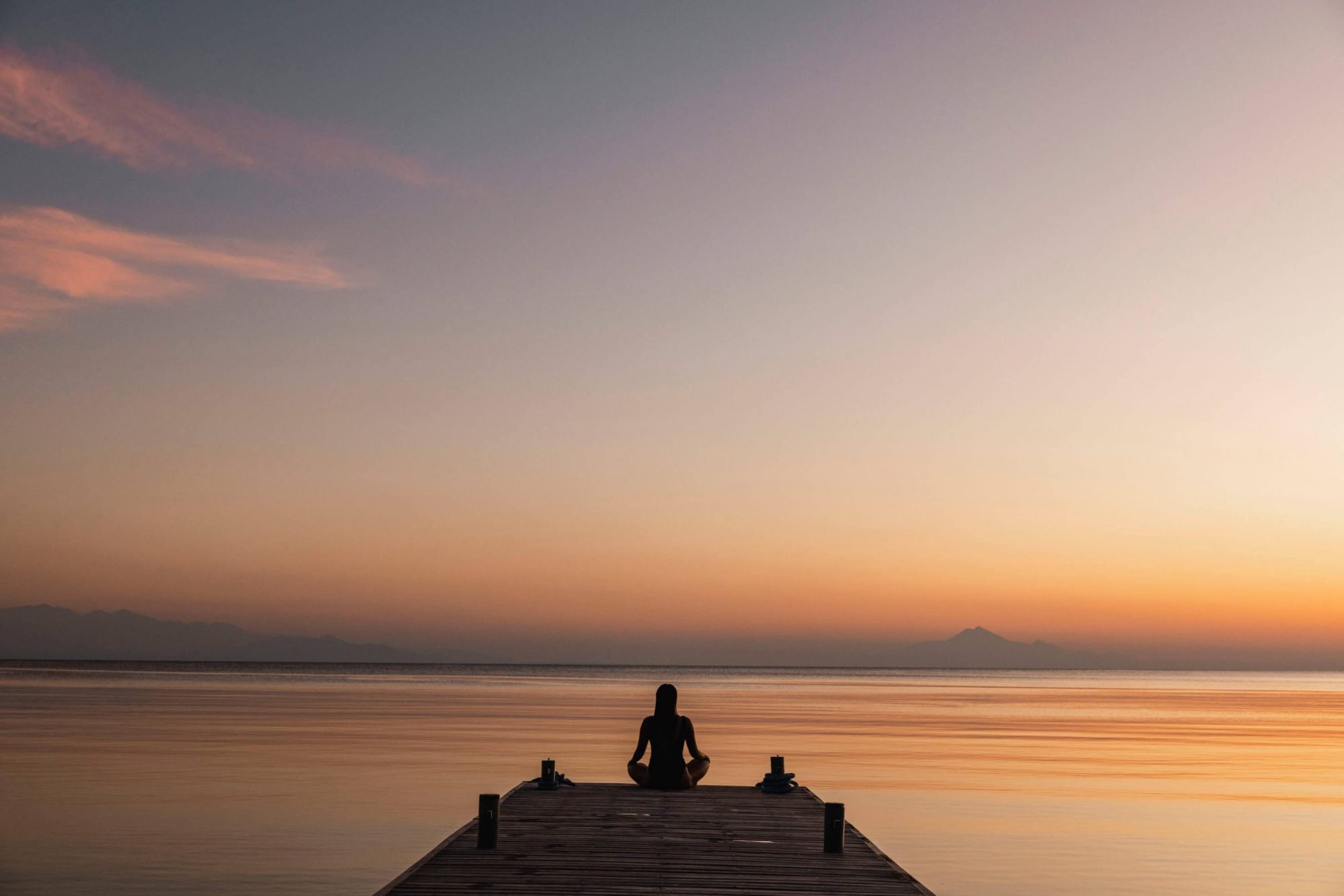 Individual meditating at sunset on a wooden dock, with calm water and a pastel sky in the background.