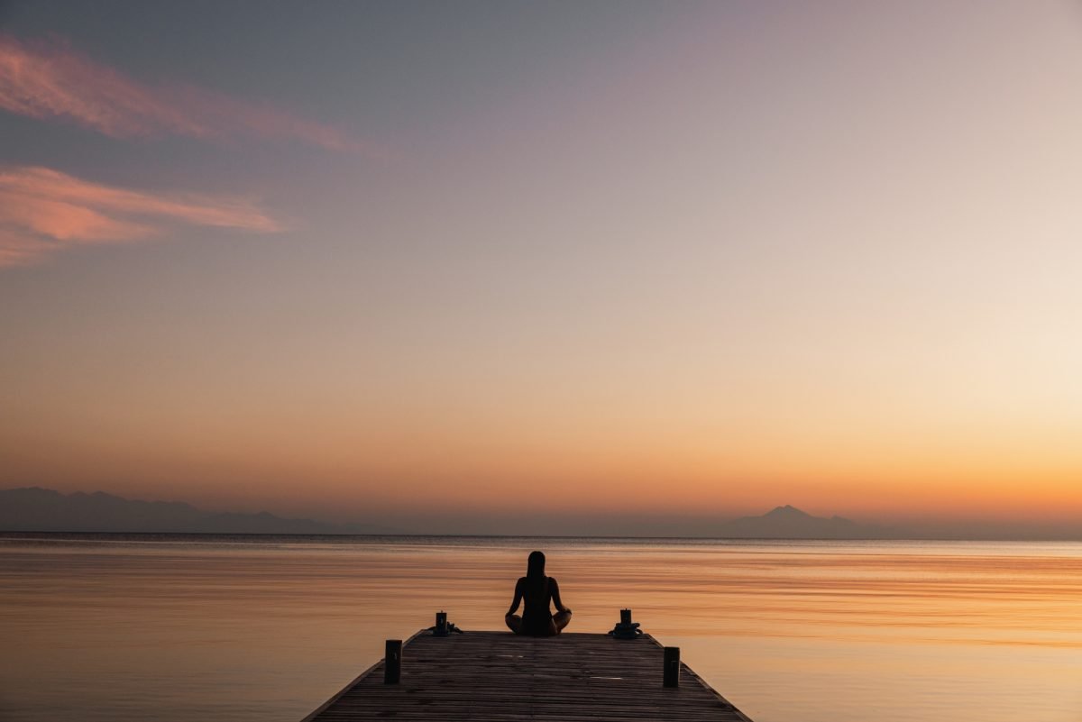 Silhouette of an individual sitting cross-legged on a wooden pier during a sunset over calm waters.