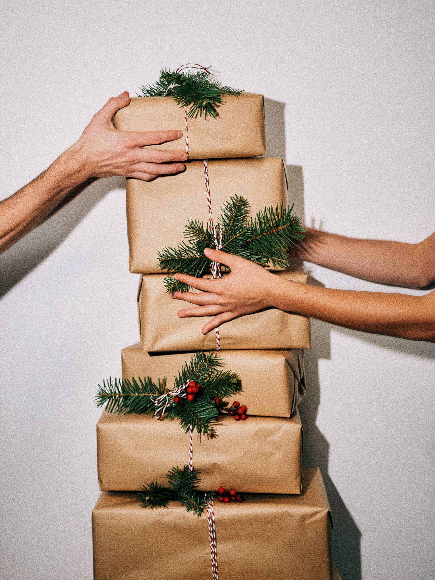 A stack of five wrapped gifts adorned with green foliage and red berries, held by two hands.