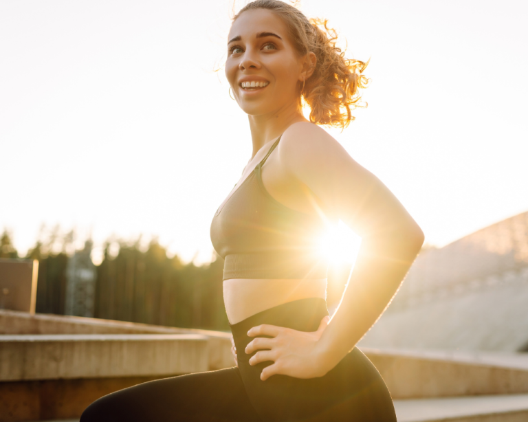 A woman in athletic wear smiling while posing outdoors with sunlight behind her.
