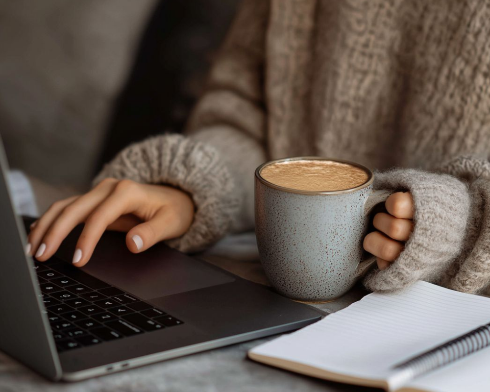Individual in a knitted sweater holding a grey mug while typing on a laptop with a notepad beside.