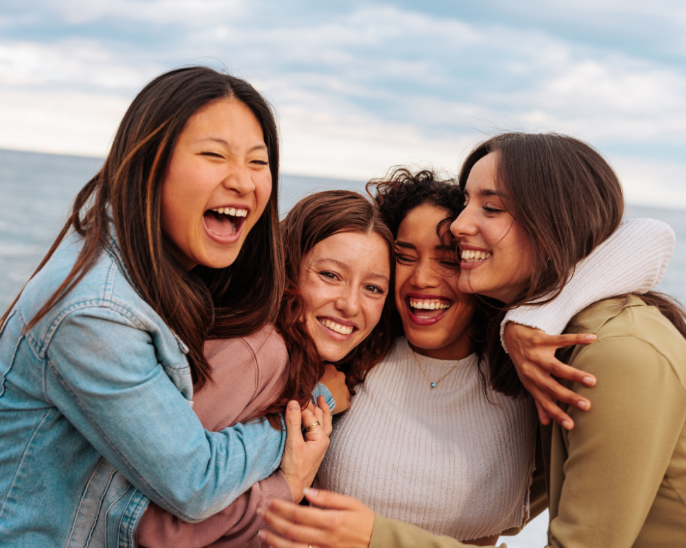 Four women laughing and embracing each other, showcasing joy and friendship by the water's edge.