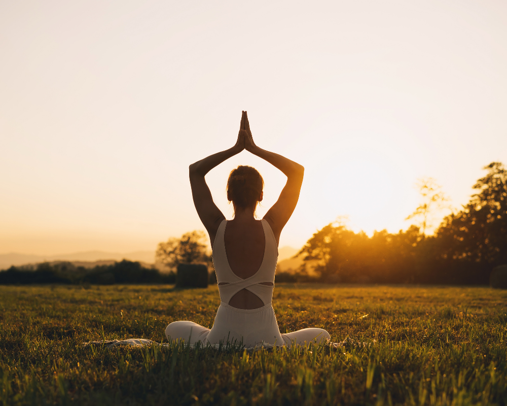 A woman in a white outfit performing a yoga pose with hands raised at sunset in a grassy field.