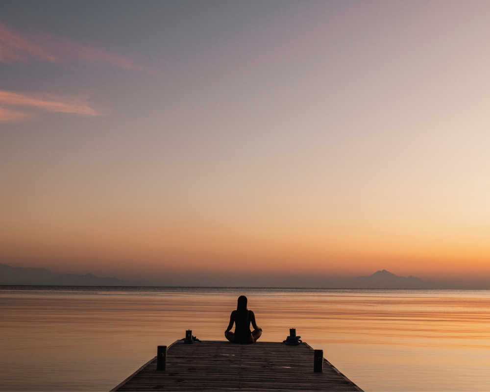 Person meditating on a wooden dock during sunset, with calm water and a serene sky in the background.