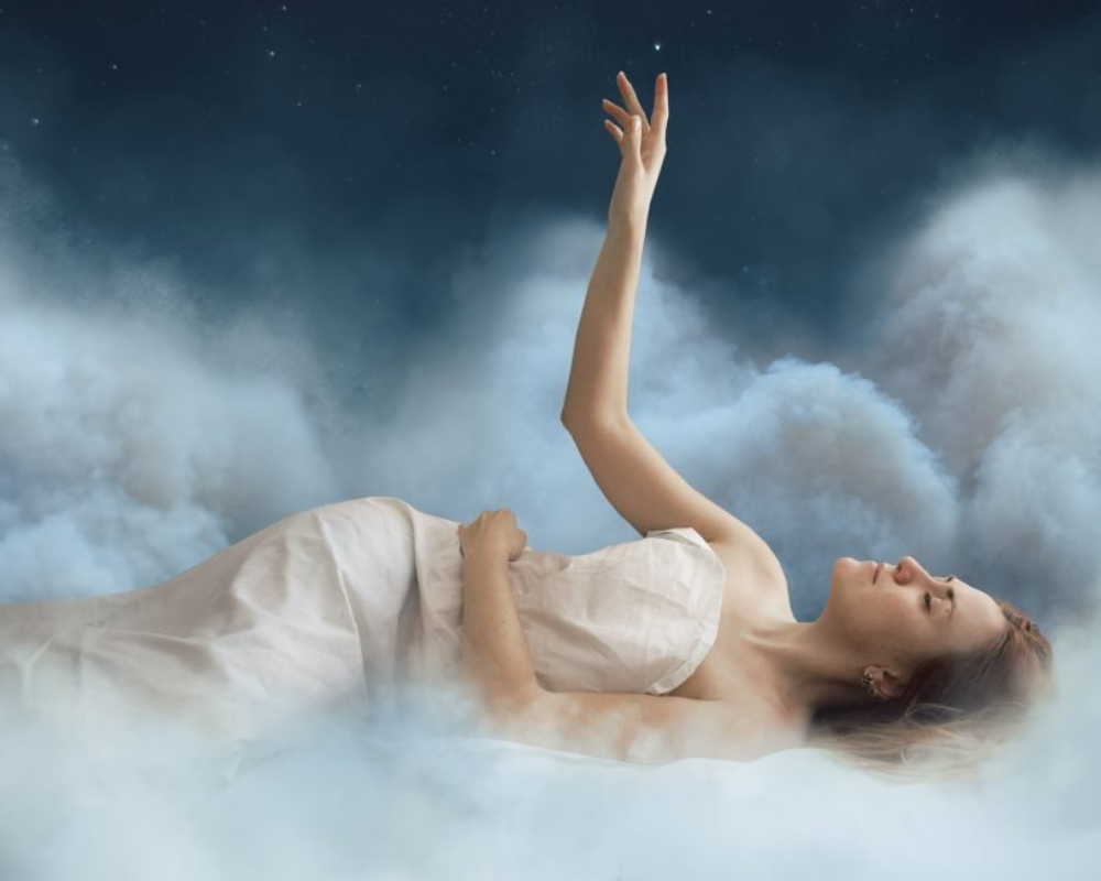 A woman lying in bed, reaching out with one hand amidst clouds in a night sky.