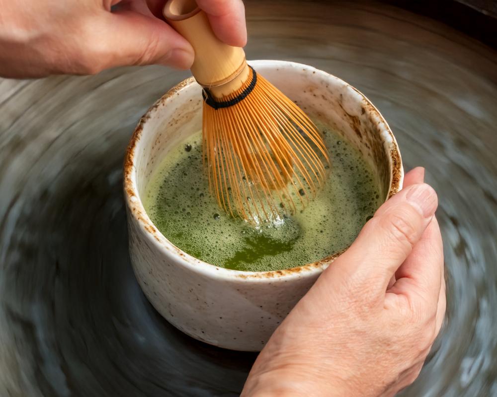 Hands using a bamboo whisk to prepare matcha tea in a ceramic bowl.