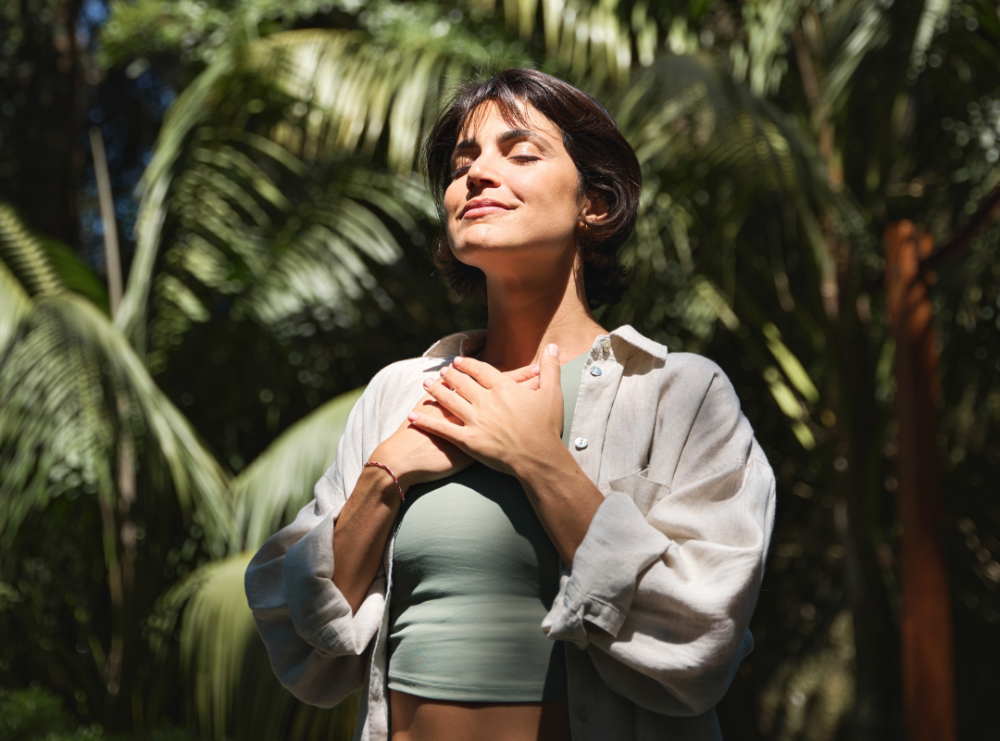 Woman standing outdoors with hands on her heart, eyes closed, and a serene expression amidst greenery.