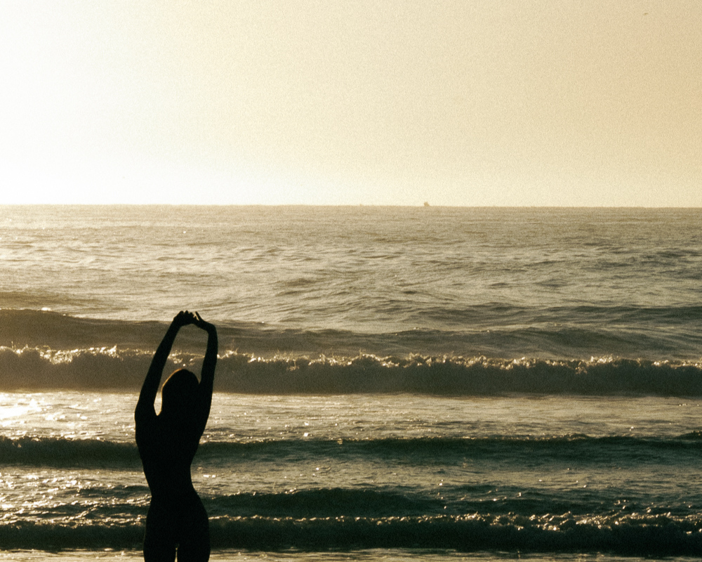 Silhouette of a person with arms raised, standing by the shoreline with waves in the background.