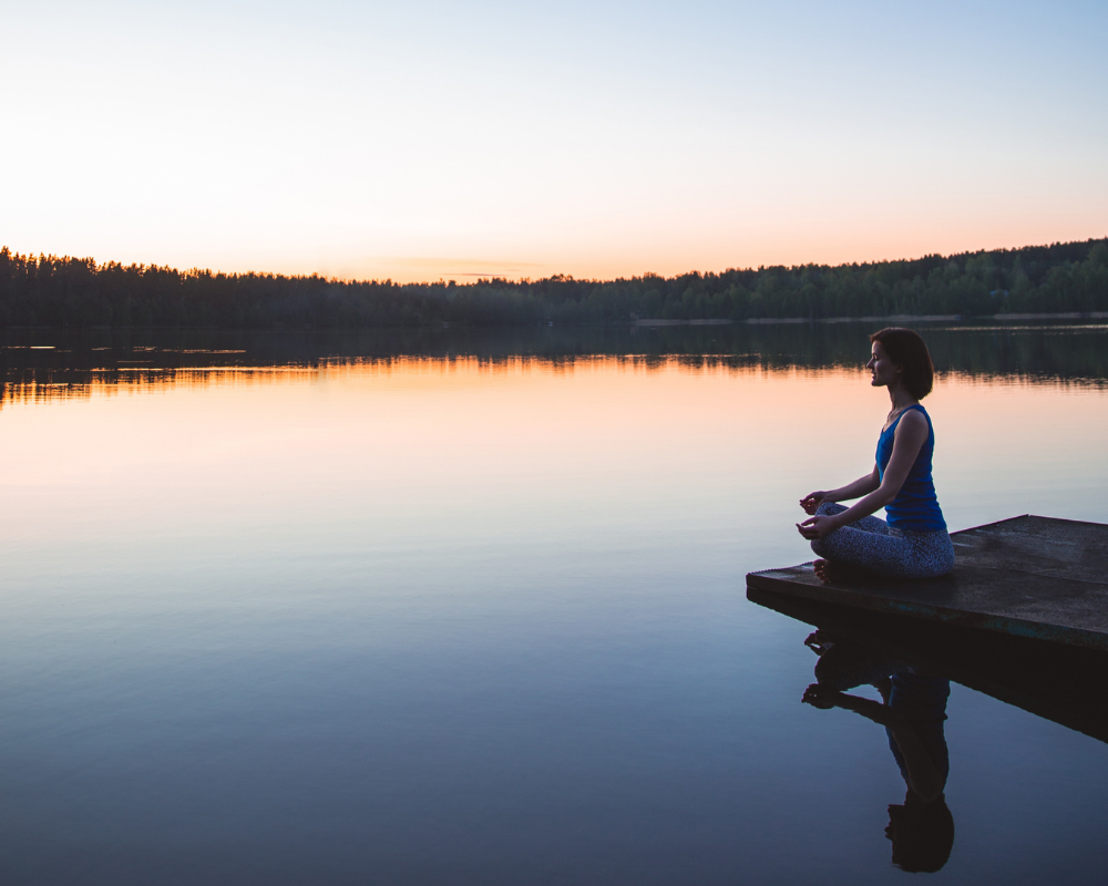 Individual meditating on a dock by a calm lake during sunset, reflecting serene colours in the water.