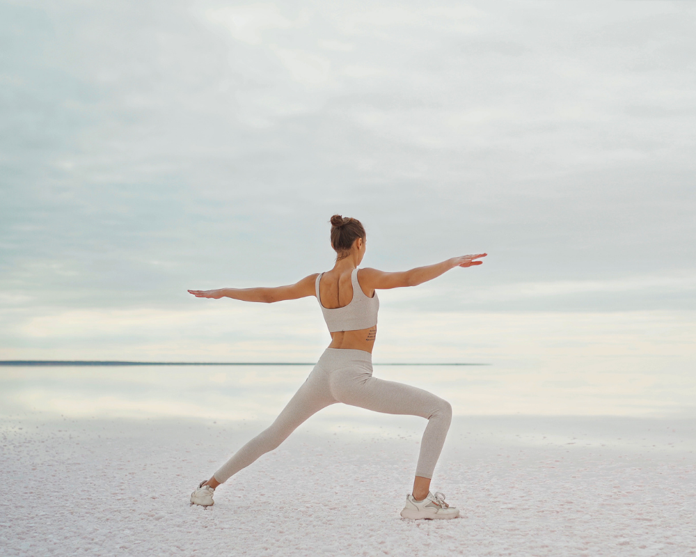 Woman in a yoga pose on a flat surface, wearing a light grey sports outfit and white trainers.