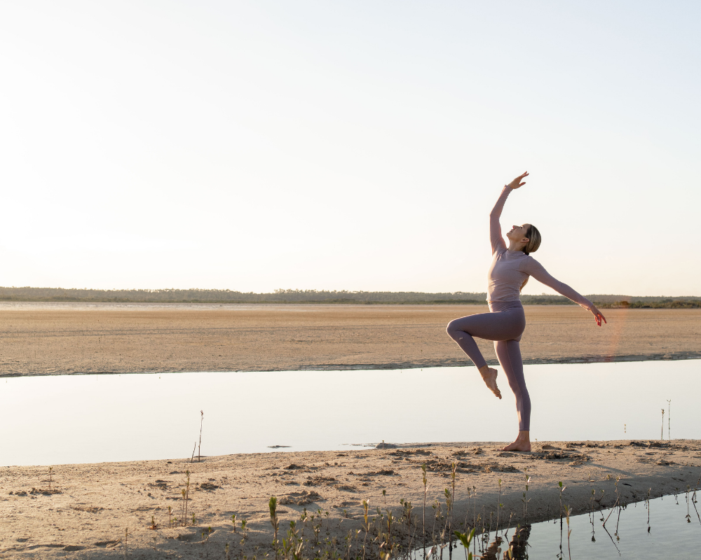 A graceful standing yoga balance at golden hour on a sandy shoreline, with calm water and soft light stretching across the horizon