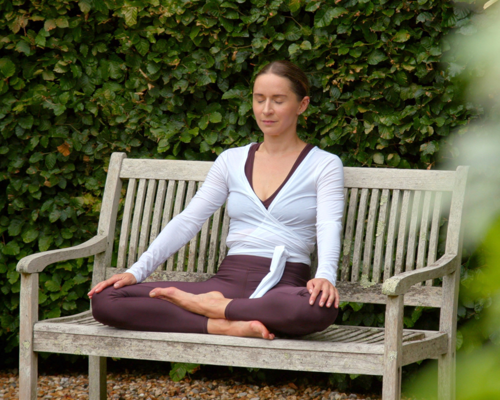 Relaxed mindfulness meditation pose on a weathered wooden bench, with a lush green hedge backdrop, white wrap top and dark maroon leggings