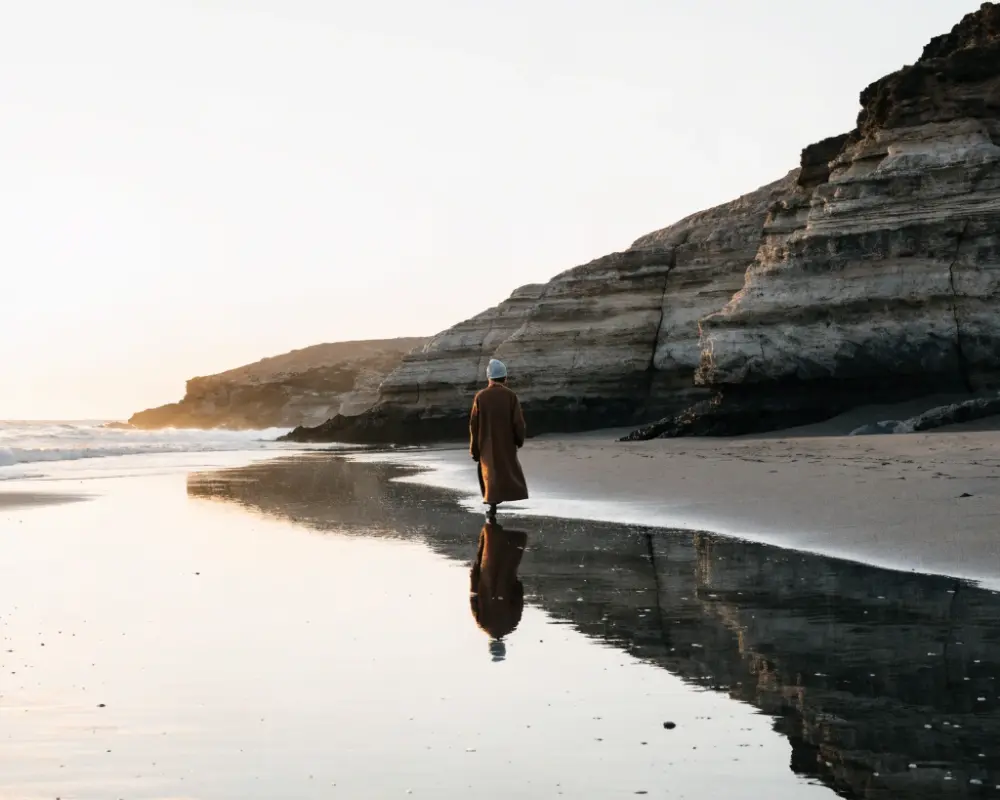 A solitary person in a brown coat and grey beanie walks along a sunlit beach, their reflection visible in a tidal pool beside rugged sandstone cliffs