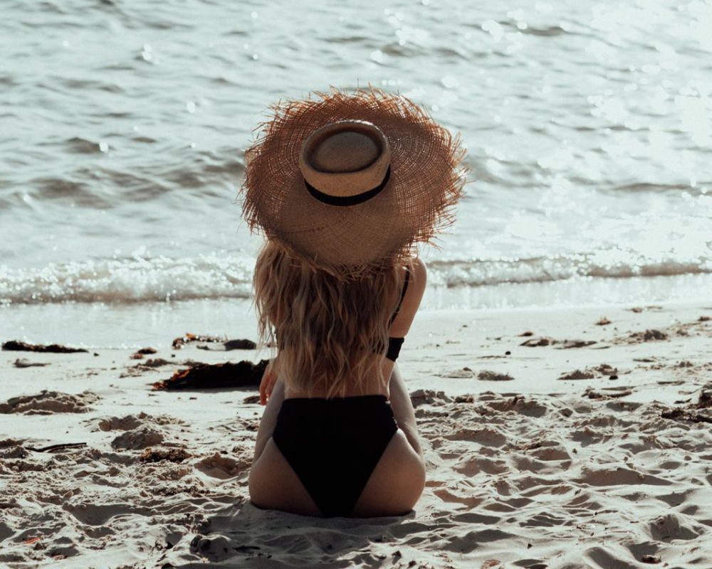 Lying on sandy beach with ocean waves behind, wearing a straw hat and a black bikini, with sunlight glinting on the water.