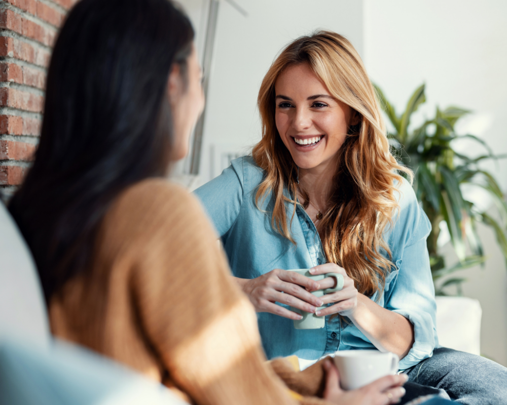 Blonde woman in a light blue shirt smiling while holding a mug, engaging in a friendly chat with a friend in a cosy setting featuring a brick wall and greenery