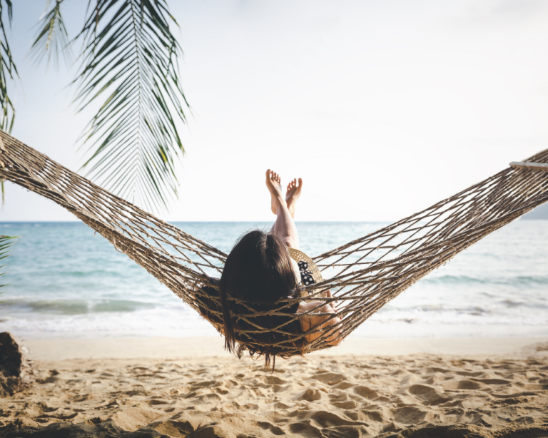 A quiet moment in a hammock over a sandy beach with palm leaves framing the view and the ocean horizon behind, reflecting rest and downshifting.
