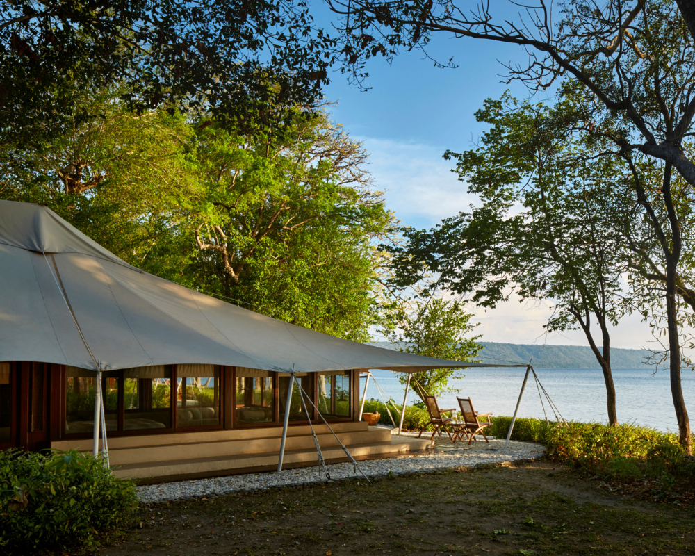 Amanwana at The Pause with an outdoor canopy beside calm lakeside water, leafy green trees framing the scene under a clear blue sky.